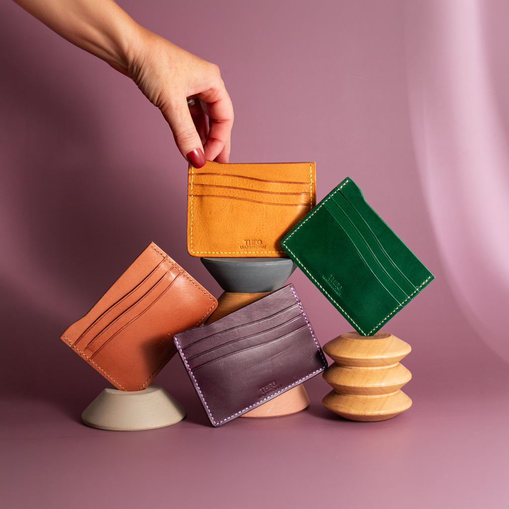 Hand holding a brown leather cardholder above four stacked cardholders on a pink background